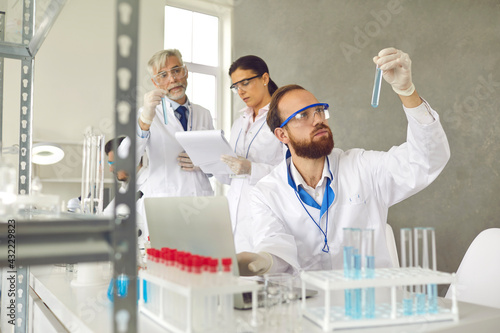 Focus on male scientist front of laptop holding test tube flask conducting chemical experiment at laboratory with senior professor and young female assistant on background. Science and research