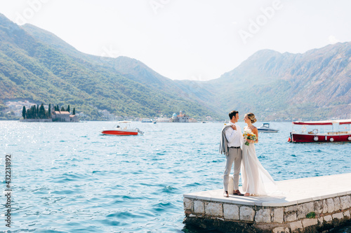  The bride and groom standing side by side on the pier in the Bay of Kotor, islands of Perast are behind them