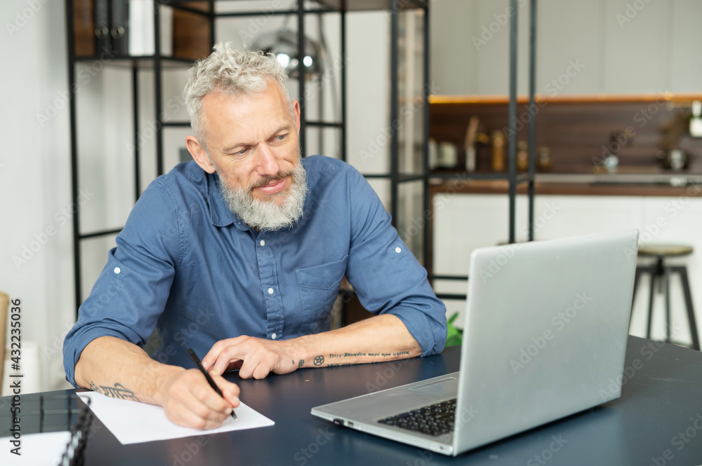 Modern middle aged bearded tattooed man working with a laptop in home office, senior grey hair male in smart casual shirt watching webinar and writes down, takes notes with a pen, planning schedule Modern middle aged bearded tattooed man working with a laptop in home office, senior grey hair male in smart casual shirt watching webinar and writes down, takes notes with a pen, planning schedule