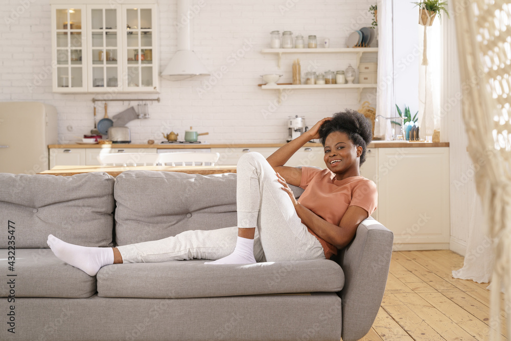 Happy smiling black woman relaxing on sofa in living room. African american female confident and relaxed look in camera with toothy smile day dreaming having rest after work at home during covid-19 Happy smiling black woman relaxing on sofa in living room. African american female confident and relaxed look in camera with toothy smile day dreaming having rest after work at home during covid-19