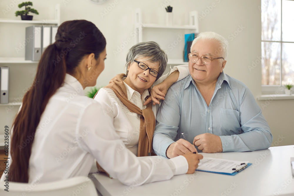 Smiling senior couple talking to nurse or their family practitioner sitting at table at doctor's office. Happy mature patients listening to physician who's explaining prescribed course of treatment Smiling senior couple talking to nurse or their family practitioner sitting at table at doctor's office. Happy mature patients listening to physician who's explaining prescribed course of treatment