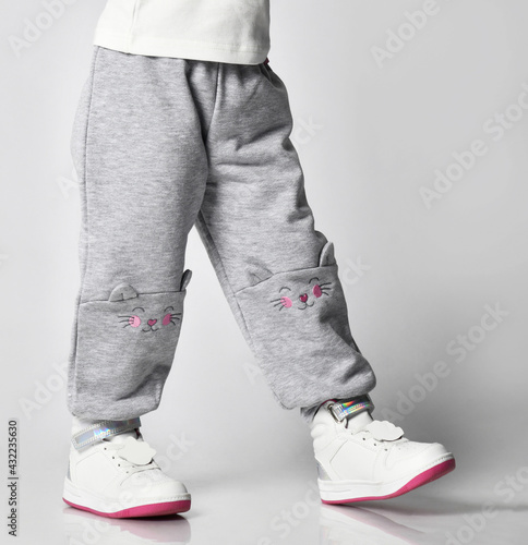 Close-up of a child in gray sports pants with a print of a cat s face and white sneakers on a gray background. Foreground. Childhood  fashion  advertising. Full length  copy space