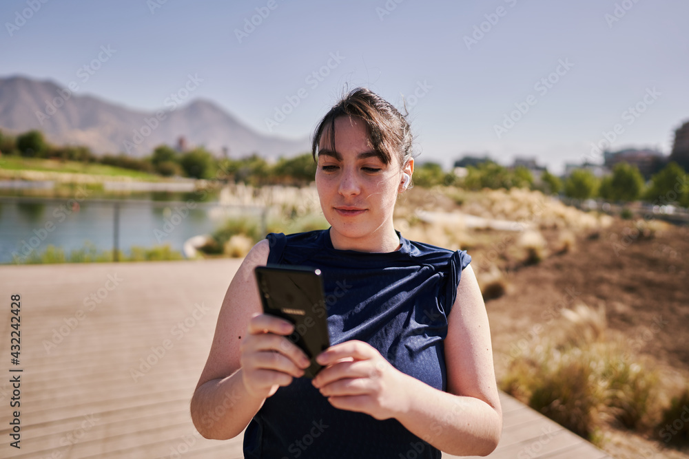 A beautiful white woman with brown hair and a sports suit takes a selfie exercising and listening to music in a park in the city center