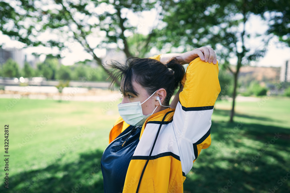 Naklejka premium A beautiful white woman with brown hair and a sports suit with a yellow jacket prepares for exercise and exercises her muscles to avoid cramps in a park near the city wearing a Covid 19 mask 