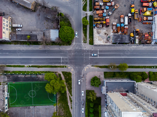 City road intersection with no humans, three cars and football court near the road. Road pedestrian crosswalk with park zone and junkyard nearby