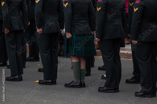 A group of men standing at attention in black suits and a kilt on parade. The men are wearing military dress uniforms. The footwear is black shiny boots. 