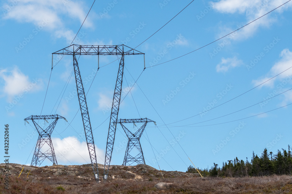 Large transmission lines and metal towers moving electricity, conductors, and distribution wires for high voltage electricity. The poles have a blue sky in the background with white clouds.