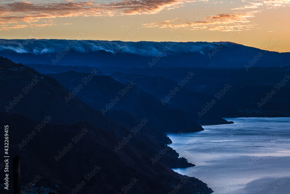 view of the lake shore surrounded by mountains in the early hours of the morning with a beautiful cloudscape with warm tones