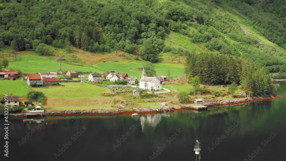 Aerial view of the village of in Bakka, Norway. A small village sitting on the coast. Green farm fields and steep, forest-covered mountains. Small tidy houses and a white wooden church. Aerial view of the village of in Bakka, Norway. A small village sitting on the coast. Green farm fields and steep, forest-covered mountains. Small tidy houses and a white wooden church.