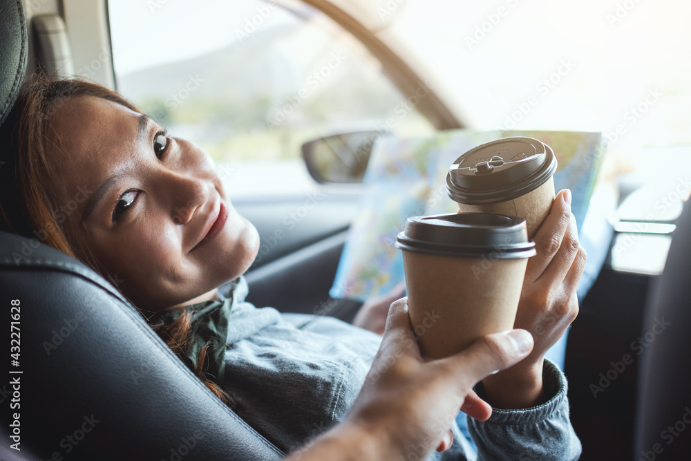 A young woman holding a map and clinking coffee cup with friend while riding in the car - obrazy, fototapety, plakaty 