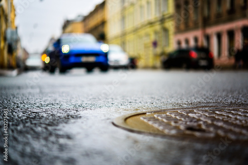 Rainy day in the big city, the headlights of the approaching blue car on the road. Close up view of a hatch at the level of the asphalt