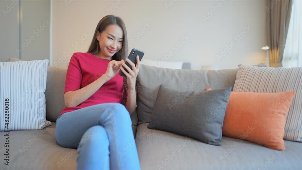 Asian woman playing mobile game on her smartphone sitting on comfortable coach with many pillow at home. Asian girl using cellphone to chat with friends Asian woman playing mobile game on her smartphone sitting on comfortable coach with many pillow at home. Asian girl using cellphone to chat with friends