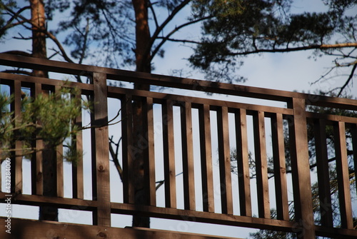 Wooden hedge railing against the sky. Railings made of dark wood against the background of a light blue sky and growing tall pines.