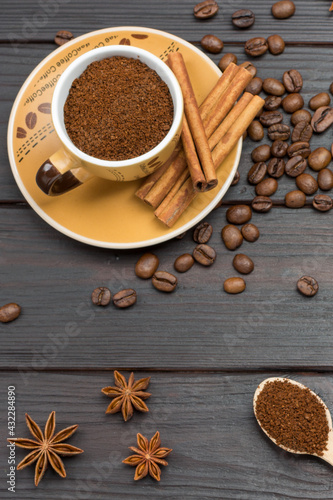 Ground coffee in cup and in wooden spoon. Cinnamon sticks on saucer. Coffee beans and star anise on table