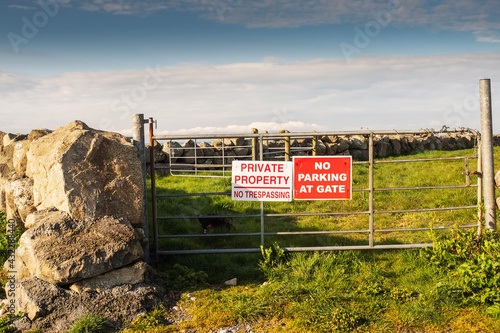 Private property no trespassing and no parking at gate signs on a metal gate. Entrance to a agriculture land. Stone fence and blue cloudy sky in the background.