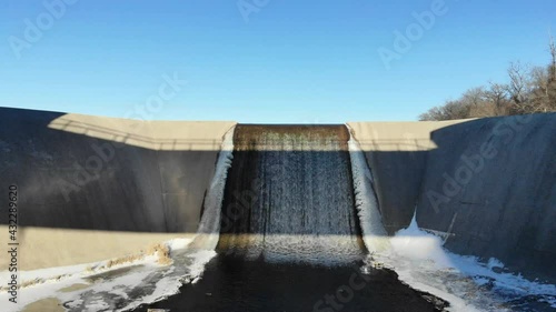 Drone flying under a bridge up and over a dam spillway out across a partially frozen lake in winter photo