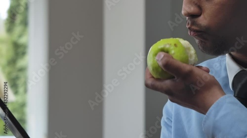 Close-up of tattooed unrecognizable African American man eating healthful apple looking at tablet screen. Serious young confident businessman working online on coronavirus pandemic in home office
