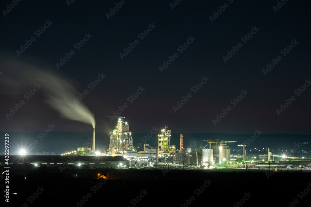 High concrete structure of cement factory with tower crane and smoking chimney at night. Industrial production and air pollution concept.