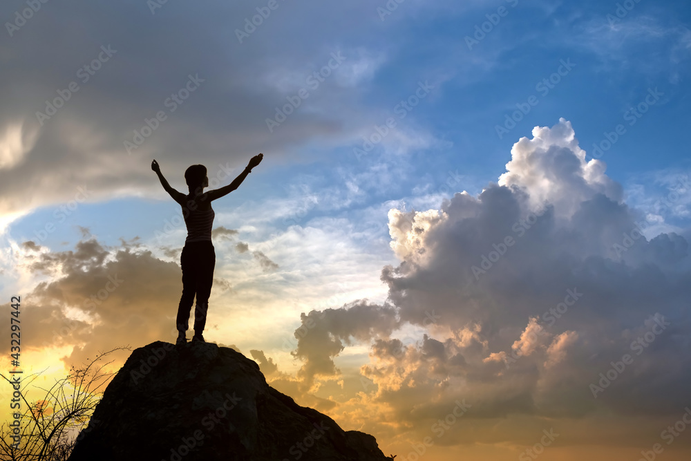 Silhouette of a woman hiker standing alone on big stone at sunset in mountains. Female tourist raising her hands up on high rock in evening nature. Tourism, traveling and healthy lifestyle concept. Silhouette of a woman hiker standing alone on big stone at sunset in mountains. Female tourist raising her hands up on high rock in evening nature. Tourism, traveling and healthy lifestyle concept.