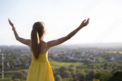Young woman in yellow summer dress standing in green meadow enjoying sunset view.