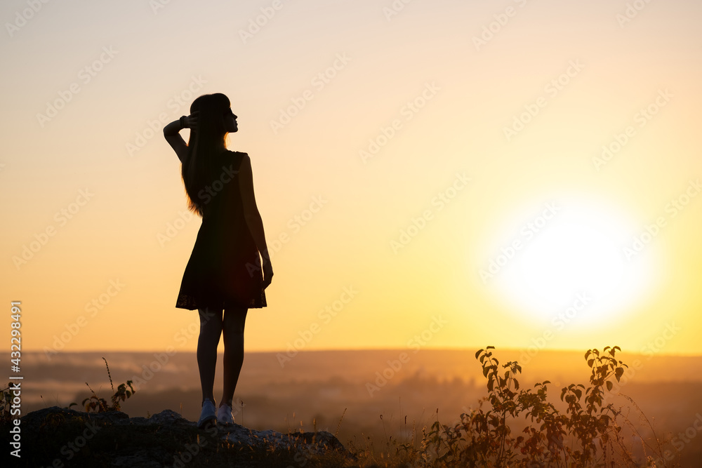 Dark silhouette of a young woman in summer dress standing outdoors enjoying view of nature at sunset.