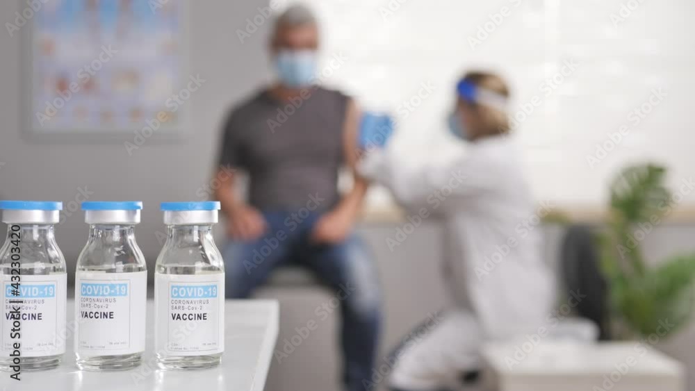 Covid-19 vaccination,close up on coronavirus vaccine bottles vials,female doctor vaccinates mature man in the background out of focus,hospital clinic studio interiors Covid-19 vaccination,close up on coronavirus vaccine bottles vials,female doctor vaccinates mature man in the background out of focus,hospital clinic studio interiors
