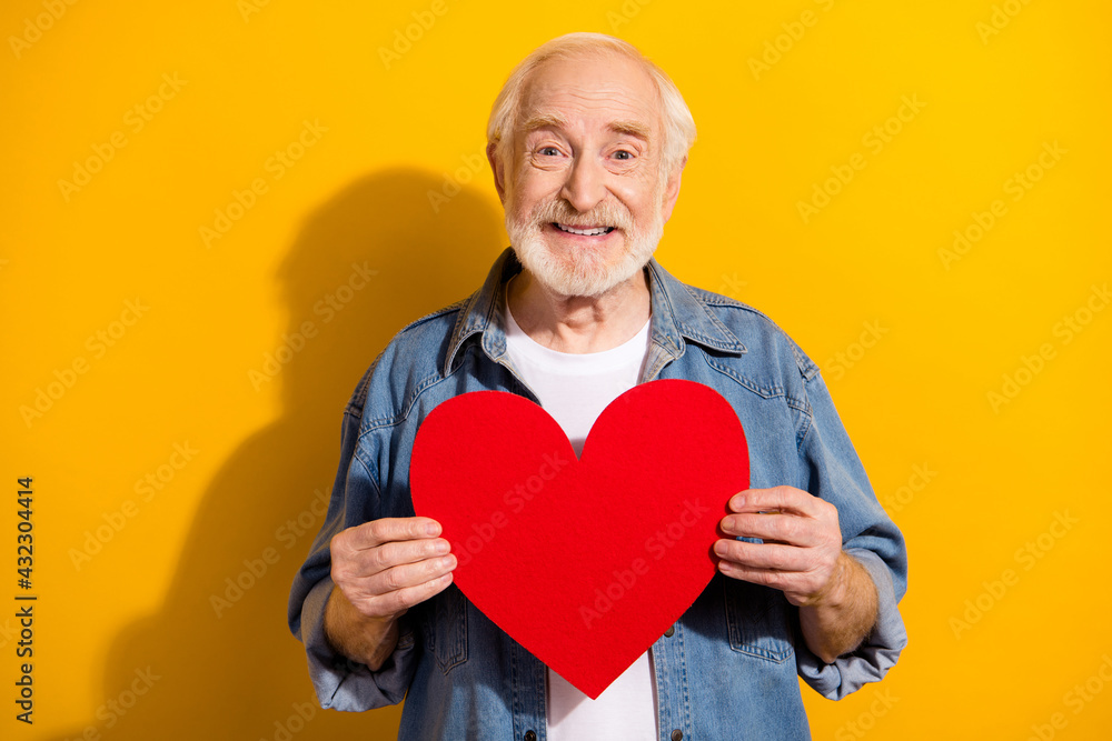 Photo portrait of elder man in jeans jacket keeping red heart smiling isolated vibrant yellow color background Photo portrait of elder man in jeans jacket keeping red heart smiling isolated vibrant yellow color background