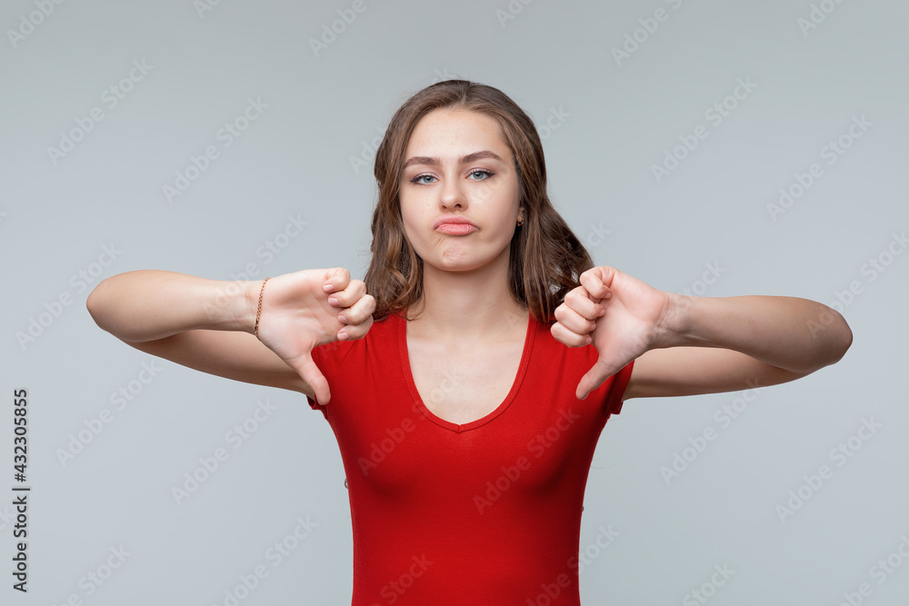 Portrait of a cute young brunette woman in red casual t shirt showing thumbs down. Gray background