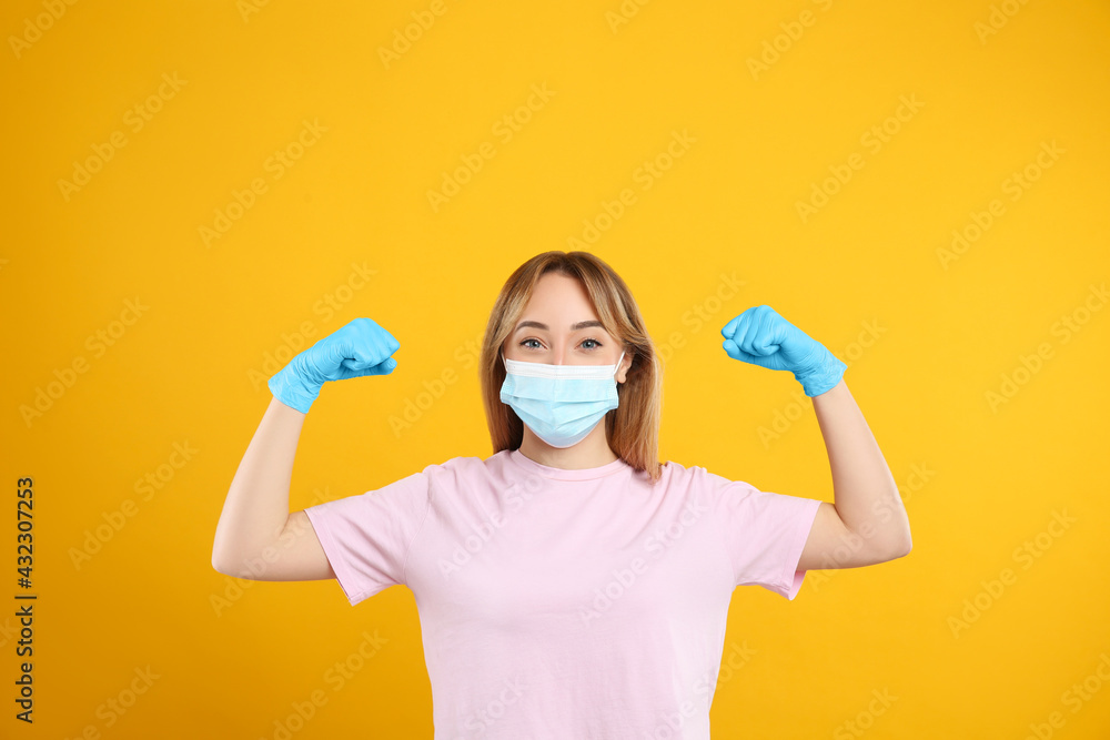 Woman with protective mask and gloves showing muscles on yellow background. Strong immunity concept Woman with protective mask and gloves showing muscles on yellow background. Strong immunity concept