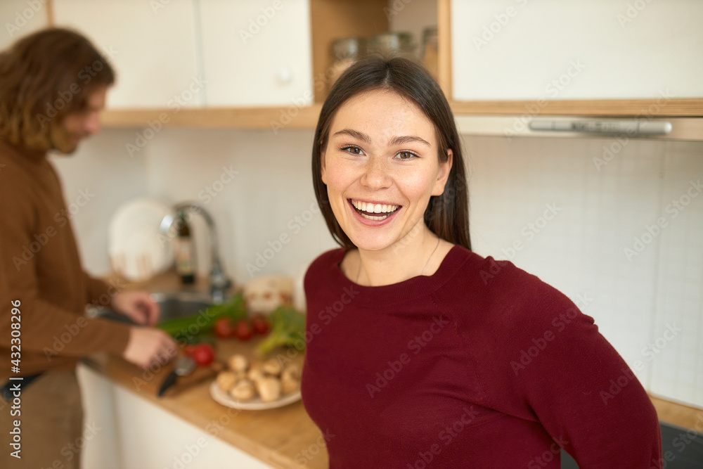 Cheerful young female wearing casual clothing posing in cozy kitchen interior looking at camera with broad smile, her husband standing in background chopping fresh vegetables on counter for lunch Cheerful young female wearing casual clothing posing in cozy kitchen interior looking at camera with broad smile, her husband standing in background chopping fresh vegetables on counter for lunch