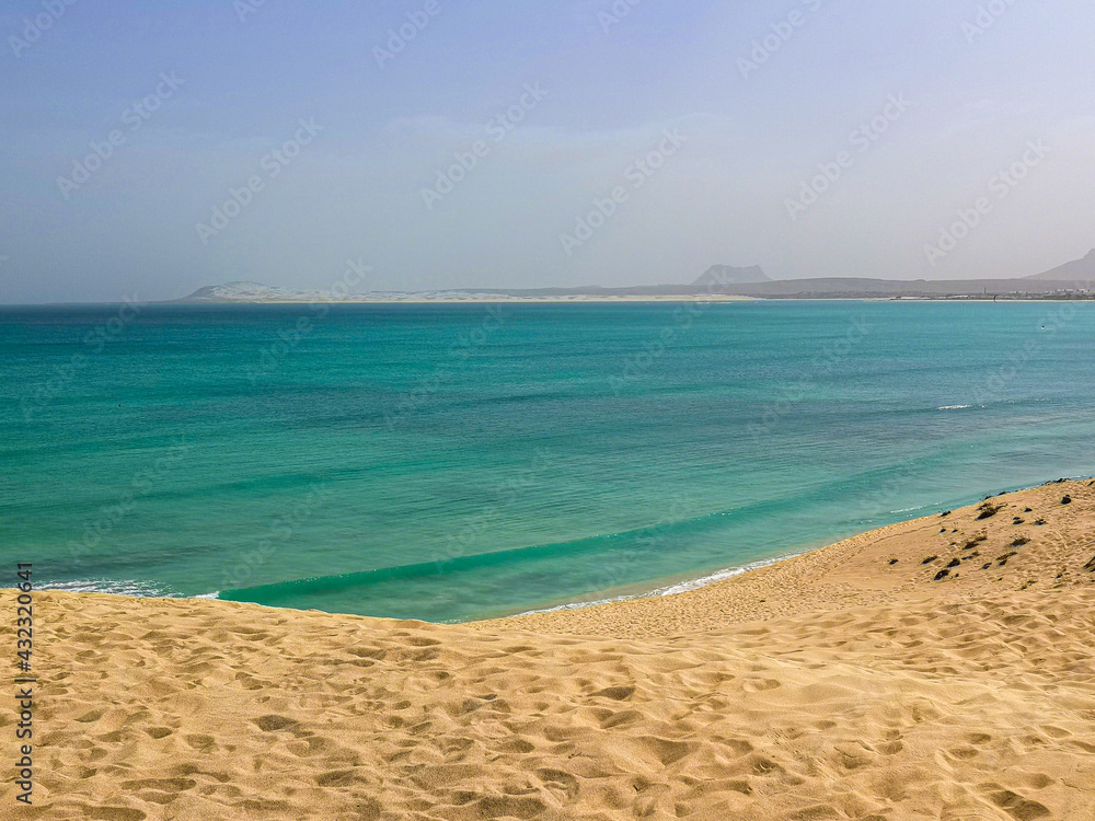 Atlantic Ocean view from a large sand dune, Boa Vista Island, Cape Verde. Clear blue water and a hot tropical day in Africa. Selective focus on the footprint pattern, blurred background.