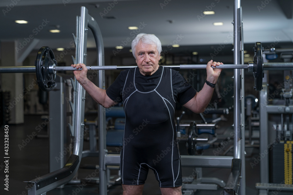 an elderly man working out in the gym stands with a barbell on his shoulders an elderly man working out in the gym stands with a barbell on his shoulders