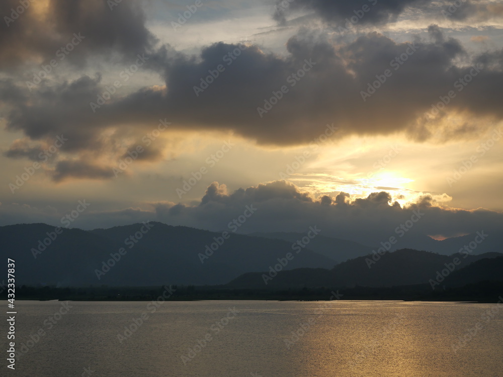 The yellow sun behind the silhouette of the cloud and mountain is reflected on the lake surface at sunset, The surface of the water sparkles with dazzling light