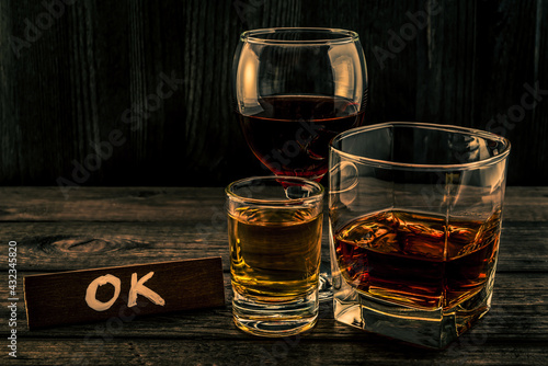 Three glasses with brandy, tequila and red wine with the wooden plank on it is an inscription "OK" on an old wooden table. Angle view focus on the inscription