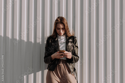 Smiling young woman in black leather jacket standing in front of white wall and using smartphone. Urban style and street fashion. Girl looking down texting on smartphone. Chatting, messaging, reading