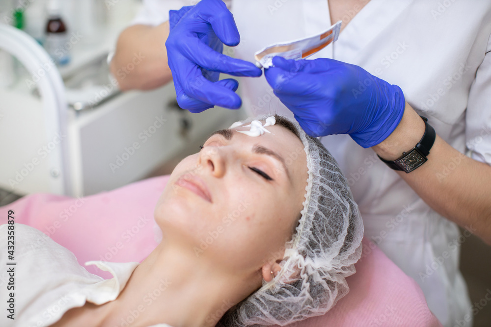 Close up top angle view of face of beautiful young woman in disposable hat, having skin cosmetology procedures in modern clinic. Hands of doctor beautician applying cream on client's face Close up top angle view of face of beautiful young woman in disposable hat, having skin cosmetology procedures in modern clinic. Hands of doctor beautician applying cream on client's face