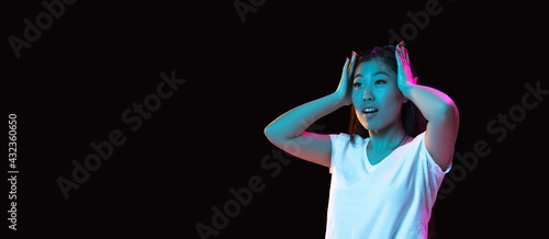 Asian young woman's portrait on dark studio background in neon. Concept of human emotions, facial expression, youth, sales, ad.