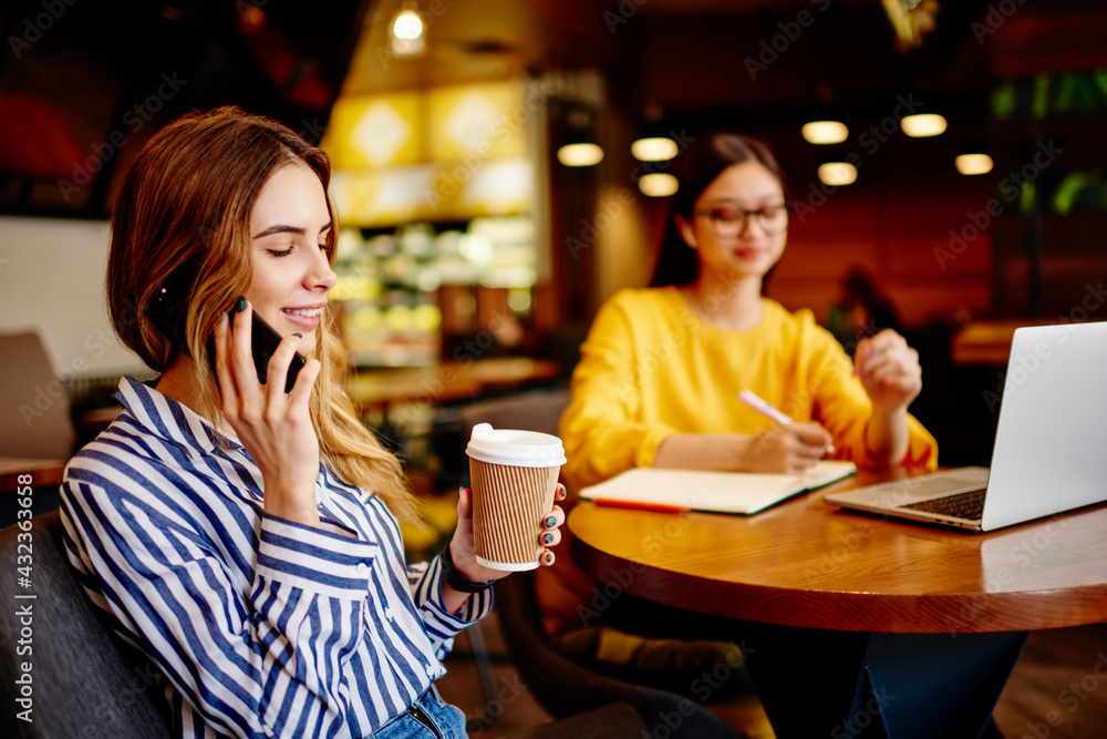 Cheerful woman speaking on smartphone and drinking coffee in cafeteria