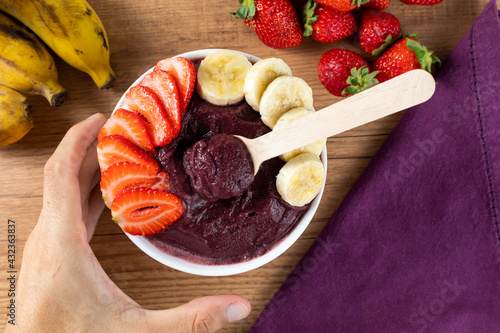 Açai, man holding an Brazilian frozen açai berry ice cream bowl. with fruits on wooden background. Summer menu front view