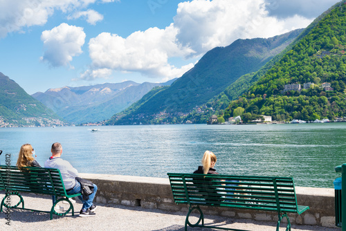 People relaxing on benches and looking the lake in a park of the city of Como, Italy. Italian alps with green forests, blue sky and white clouds on the background.