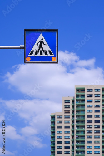 Overhead electric pedestrian crossing sign with blurred residential building on white cloud and blue sky background in vertical frame