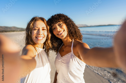 Two beautiful mixed race girl holding smartphone making a self portrait on beach smiling at camera wearing boho white dress at sunset or dawn. Gay couple of young millennial women in tropical vacation