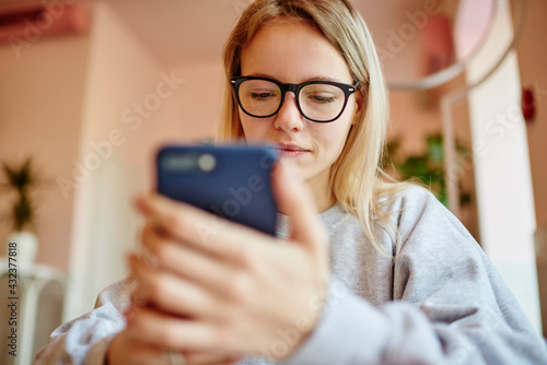 Millennial female blogger in classic eyewear browsing banking information on modern mobile phone, young Caucaisan hipster girl in spectacles installing application for making web booking on cellphone