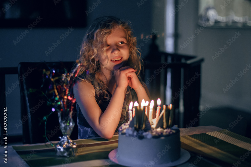 Cute funny blonde girl blowing candles on her birthday cake in dark room.  Happy birthday party celebration. Making a wish while blowing candles  tradition. Child celebrating birthday at home. Stock Photo