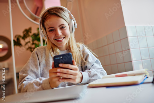 Portrait of smiling hipster girl in digital headphones for noise cancellation spending daytime for listening positive music podcast or audio book for learning, happy female blogger with mobile phone