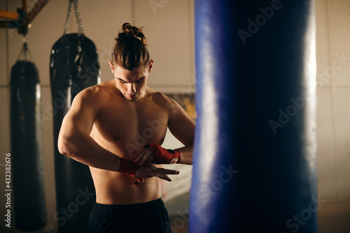 Muscular build fighter wrapping fists with boxing bandage while preparing for workout at boxing club.