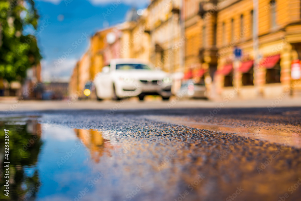 Sunny day after rain in the city, the headlights of the approaching car on the road. Close up view from the level of the puddle on the pavement