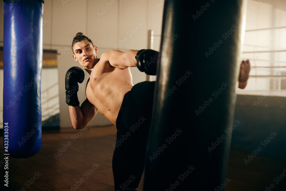 Male fighter kicking punching bag while working out at boxing club.