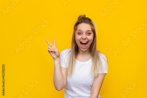 A young pretty caucasian impressed excited smiling cheerful blonde woman in a white t-shirt shows a peace gesture with her hand isolated on a bright color yellow background. Girl shows a victory sign