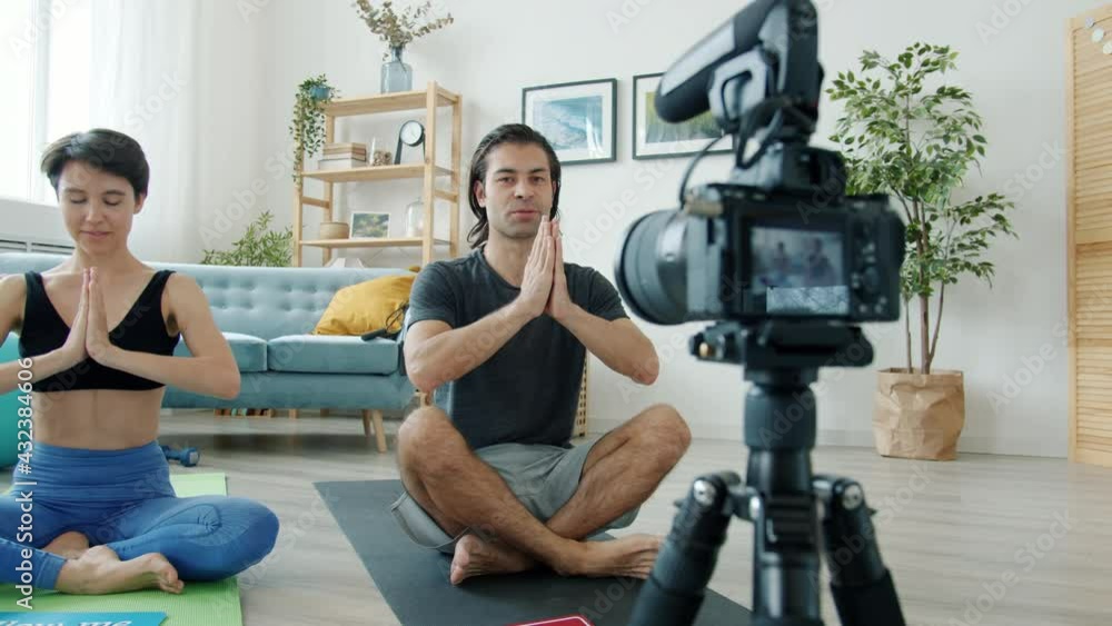 Couple of vloggers are meditating sitting on yoga mats then showing follow me sign and smiling recording video for internet blog. Vlogging and technology concept. Couple of vloggers are meditating sitting on yoga mats then showing follow me sign and smiling recording video for internet blog. Vlogging and technology concept.